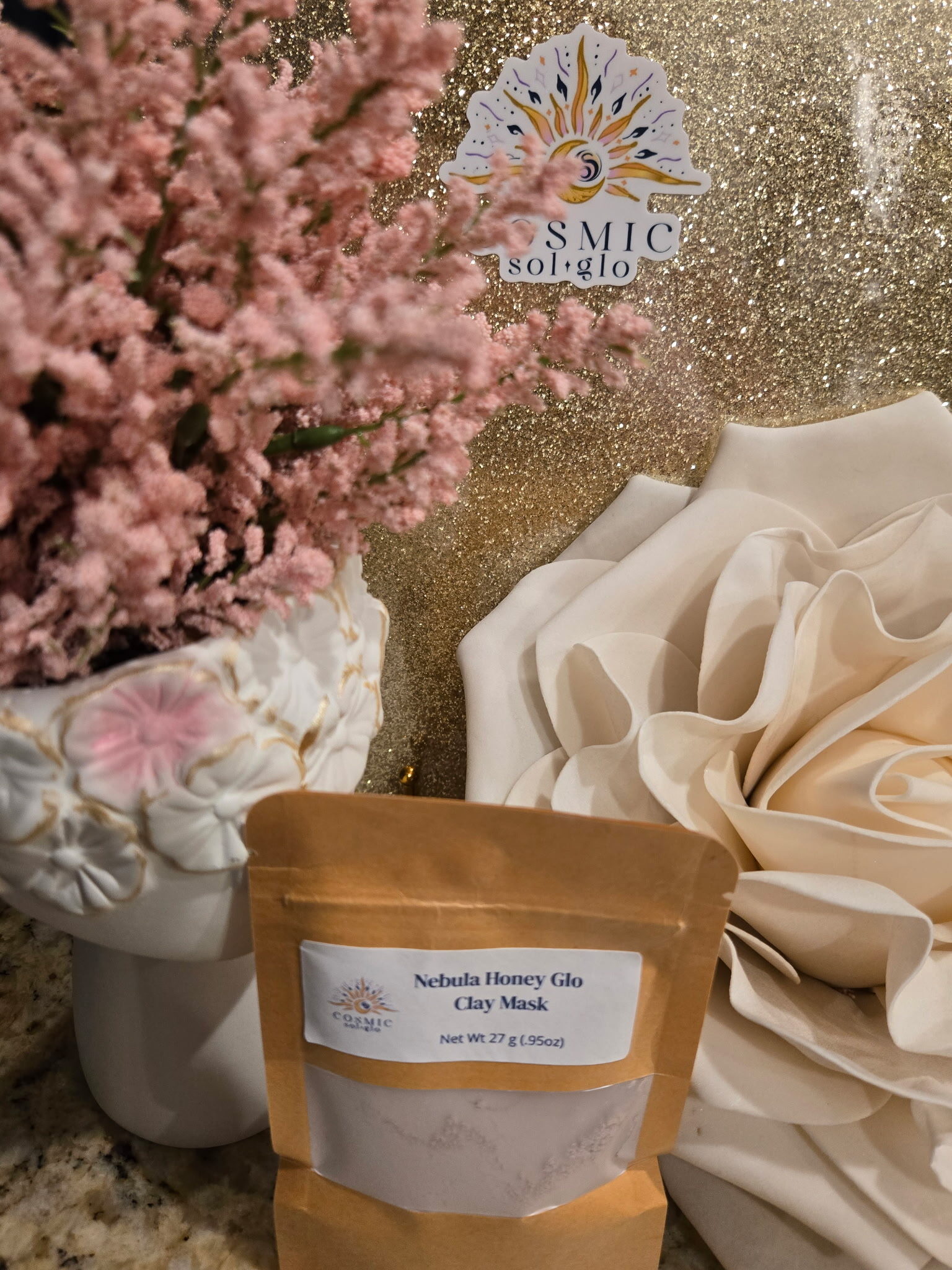 Nebula Honey Glo clay mask pouch styled with pink flowers and gold glitter